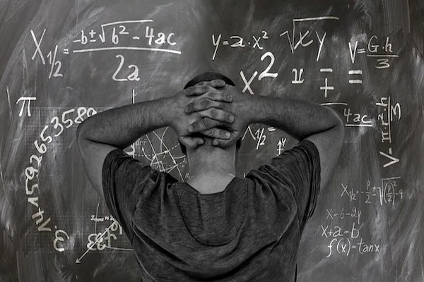 Black and white photo of male holding head in hands looking at equations on a blackboard